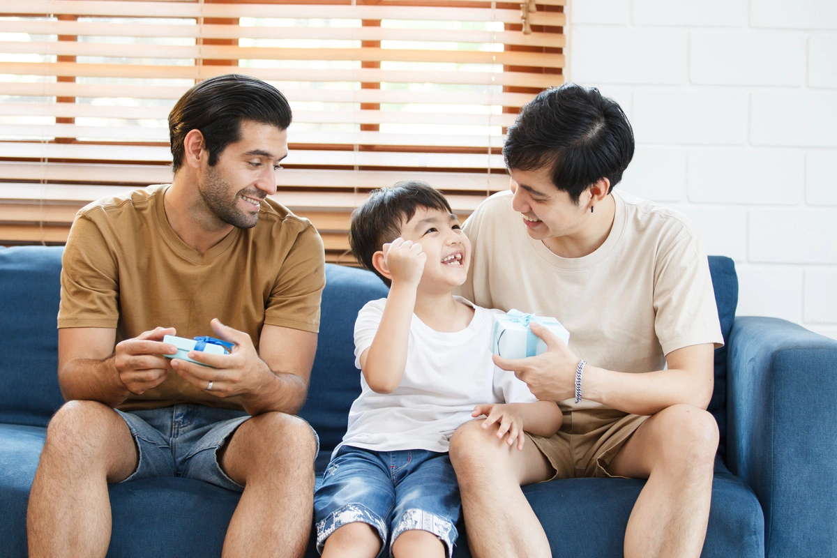 Deux hommes avec un enfant souriant tenant un cadeau, assis sur un canapé, illustrant une famille adoptive aidée par un avocat.