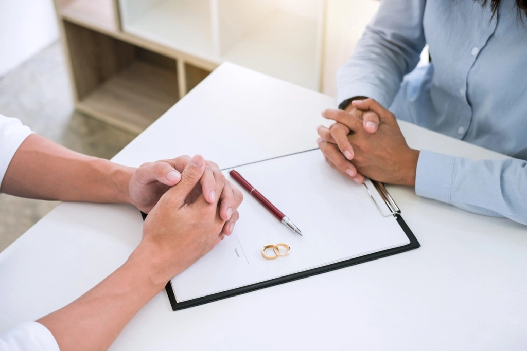 Deux personnes se faisant face, mains croisées, avec un document, un stylo et des alliances sur la table.